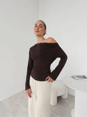 Woman wearing a brown off-shoulder top and white pants in a minimalistic room.
