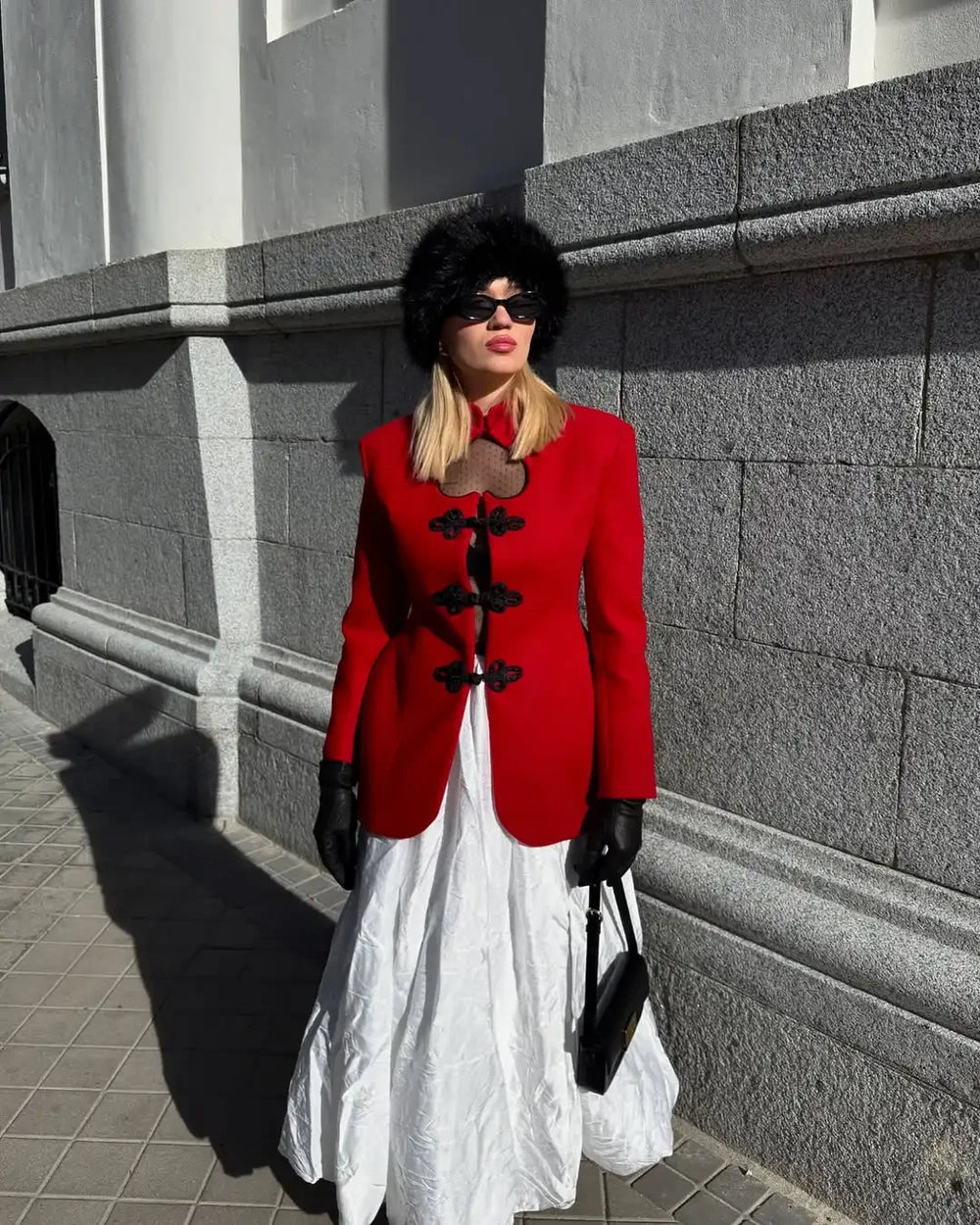 Woman in a red coat and white dress standing against a gray wall.