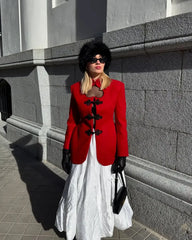 Woman in a red coat and white dress standing against a gray wall.