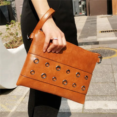 Brown leather clutch bag with grommet details held by a person on a city street.