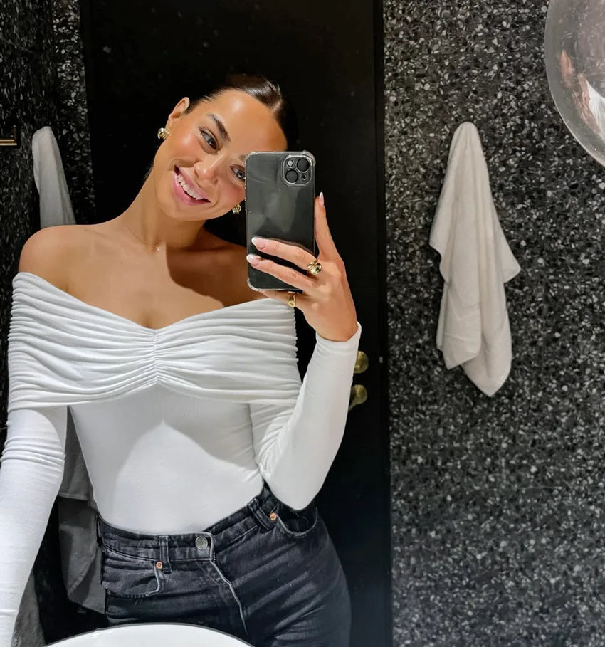 Woman taking a mirror selfie wearing a white off-shoulder top and dark jeans.