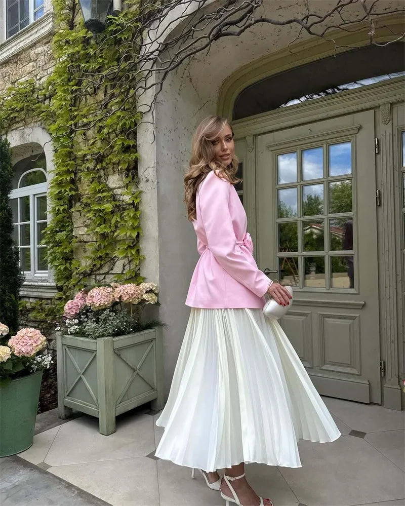 Woman in a pink jacket and white skirt standing in front of a stone building with flowers.