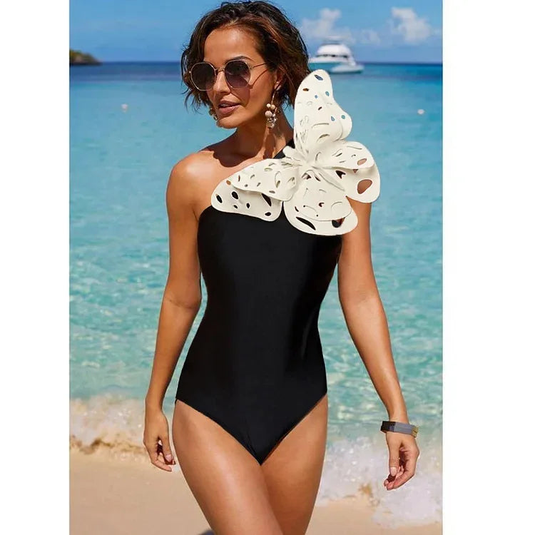 Model wearing a one-piece black swimsuit with a white 3D butterfly detail on the shoulder at the beach.