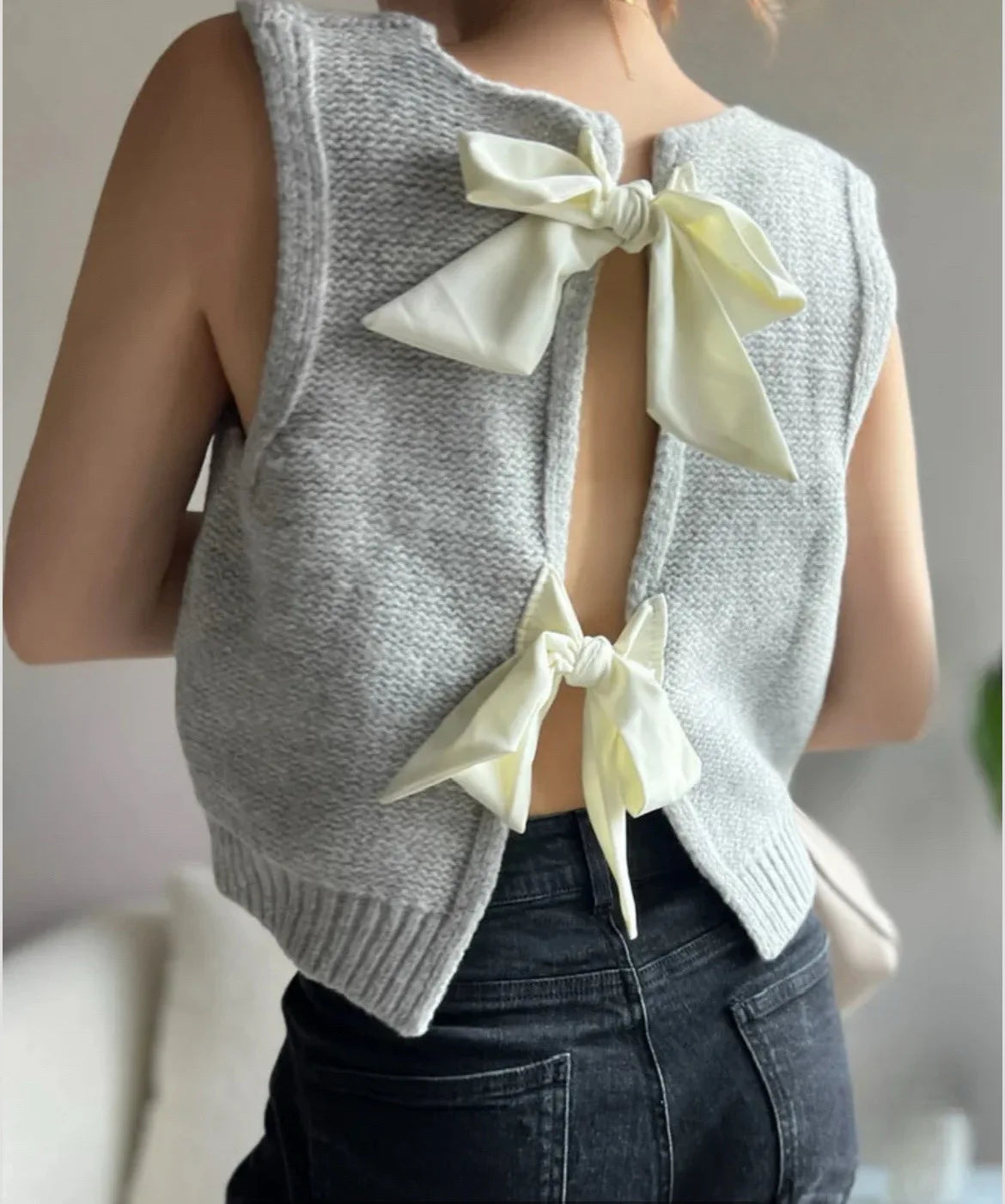 Gray sleeveless sweater with white bows worn by a person against a neutral background