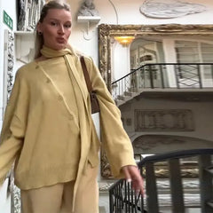 Woman in a beige outfit standing in an elegant interior setting with a staircase and mirror.