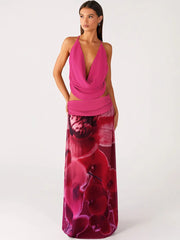 Woman in a pink cowl neck top and floral print maxi skirt posing against a white background.
