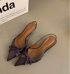 Pair of brown pointed-toe shoes with bow detail on a light gray surface, with a box labeled 'Linda' in the background.