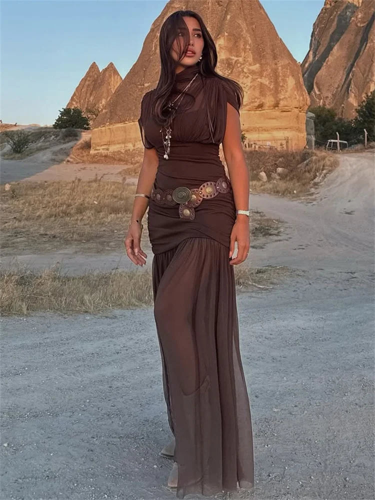 Woman in a brown dress standing in a desert landscape with rock formations.