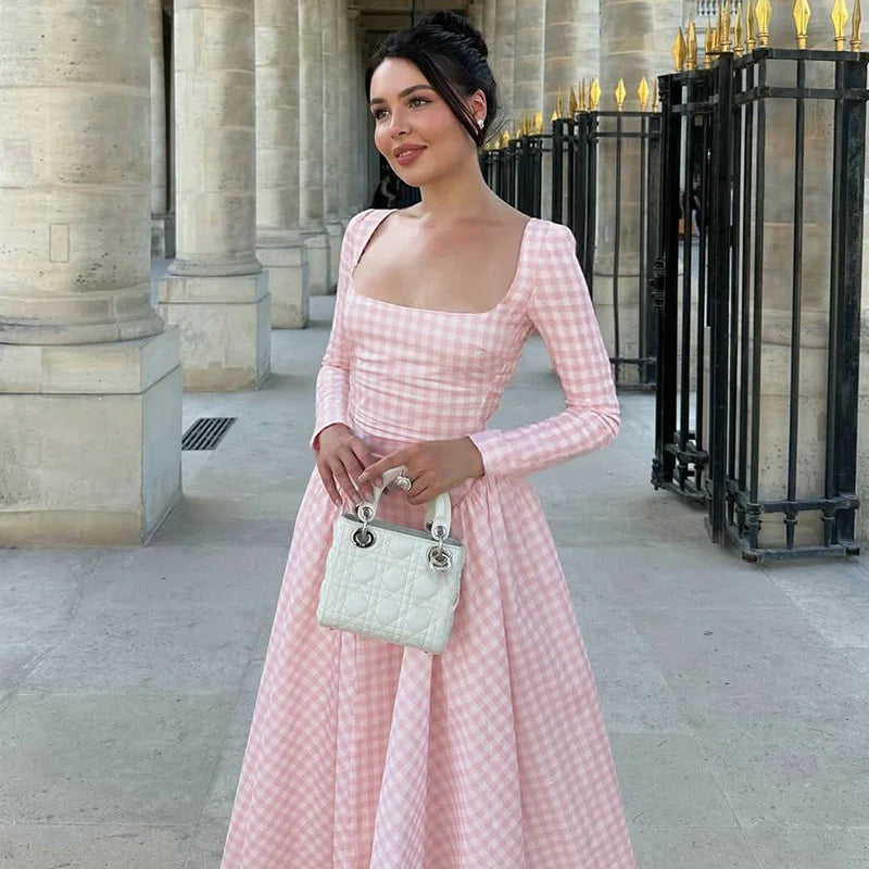 Woman in a pink checkered dress holding a white handbag outdoors.