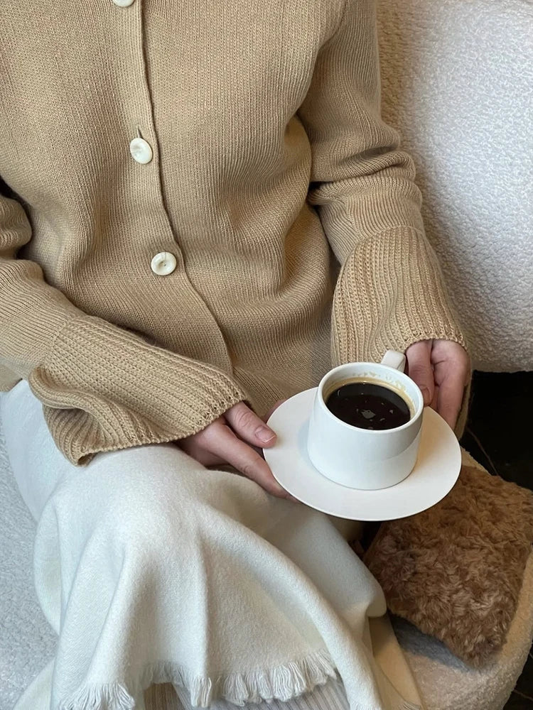 Person wearing a beige cardigan holding a white cup of coffee on a saucer.