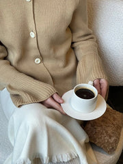 Person wearing a beige cardigan holding a white cup of coffee on a saucer.