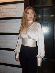 Woman wearing a white blouse with long sleeves and black pants against a textured wall.