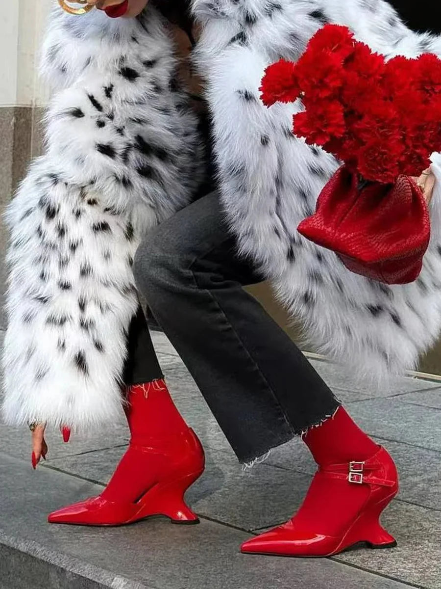 Person wearing a white fur coat with black spots, black pants, and red high-heeled shoes holding red flowers.