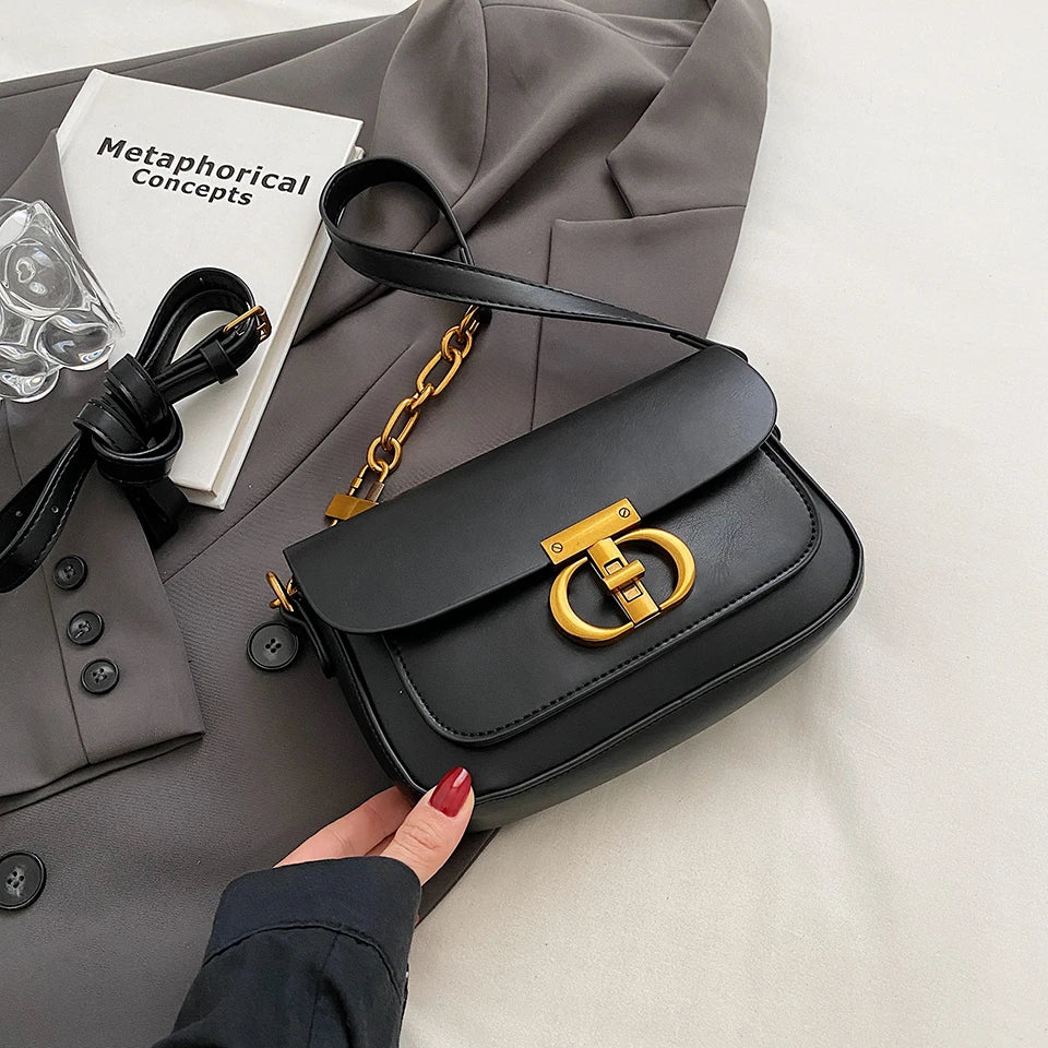 Black handbag with gold accents held by a hand on a light gray surface with a book and coat in the background.