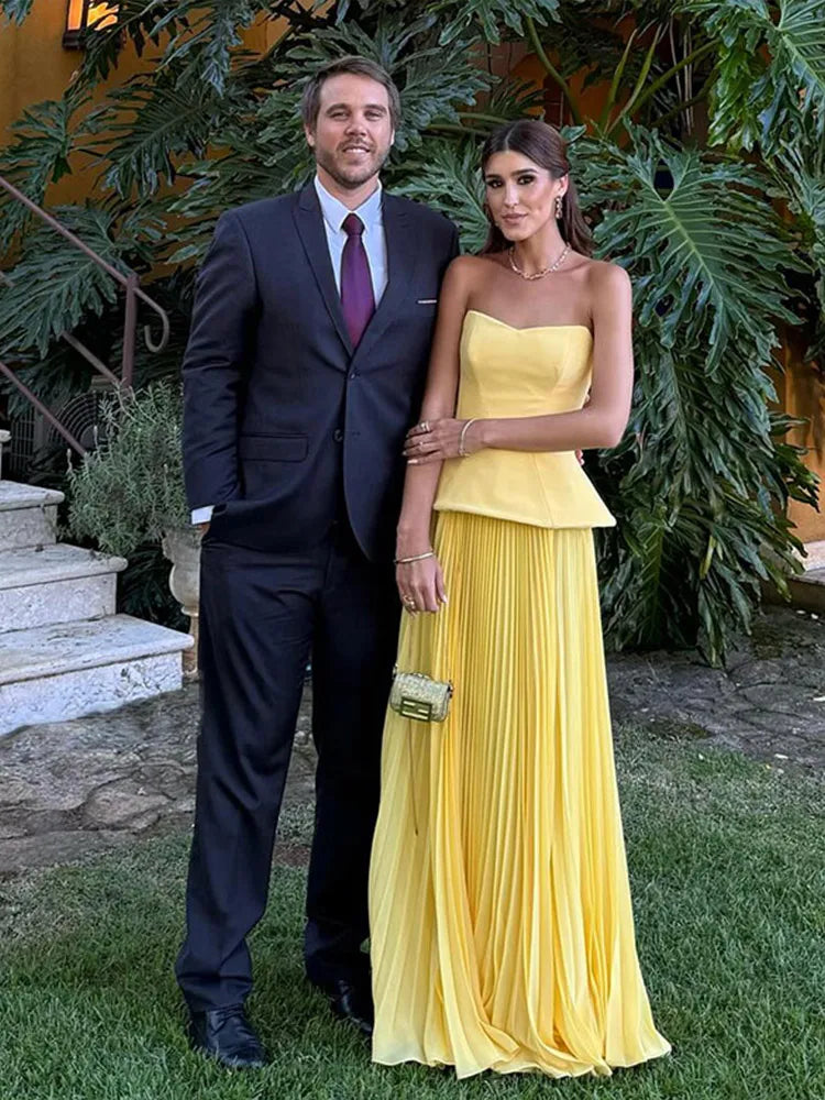 Man in a suit and woman in a yellow dress standing outdoors with greenery in the background