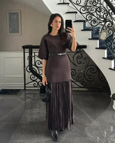 Woman taking a mirror selfie in a dark brown dress with a pleated skirt in a staircase setting.