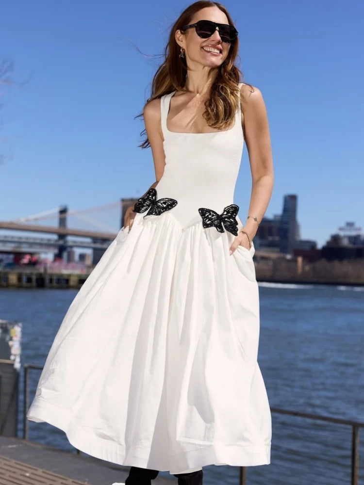 Woman in a white dress with black butterfly details standing by a waterfront with a bridge in the background.