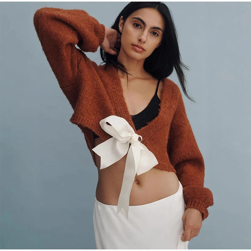 Woman wearing a brown cardigan with a white bow and white pants against a gray background