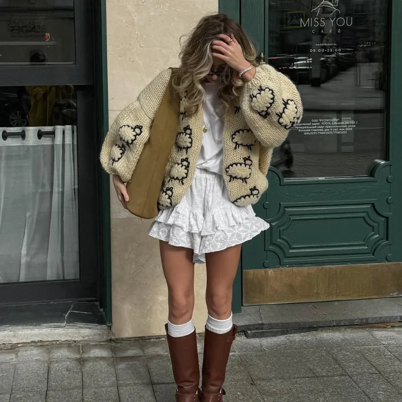 Woman wearing a patterned cardigan, white dress, and brown boots on a city street.