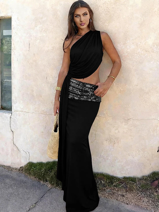 Woman in a black one-shoulder dress with a sequined waistband standing against a light-colored wall.