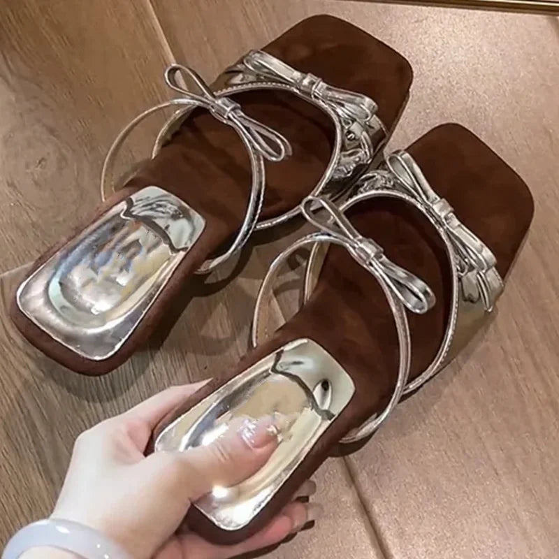 Brown sandals with silver straps held by a hand on a wooden floor.