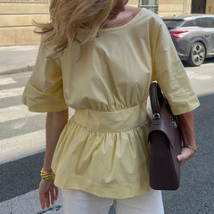 Woman wearing a yellow blouse and white pants, holding a brown bag on a street.