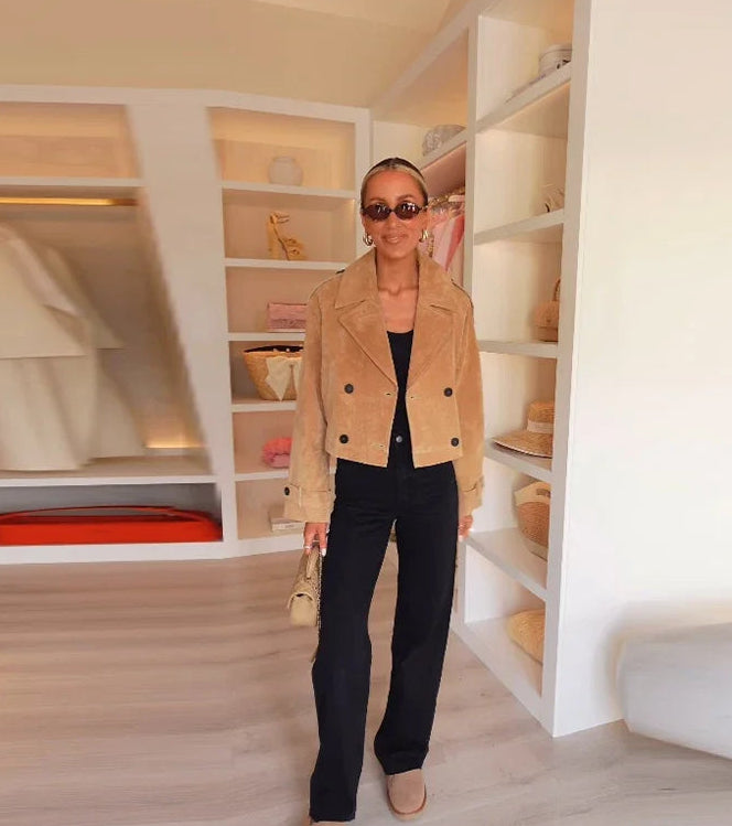 Woman in a beige jacket standing in a stylishly decorated room with shelves and decor.