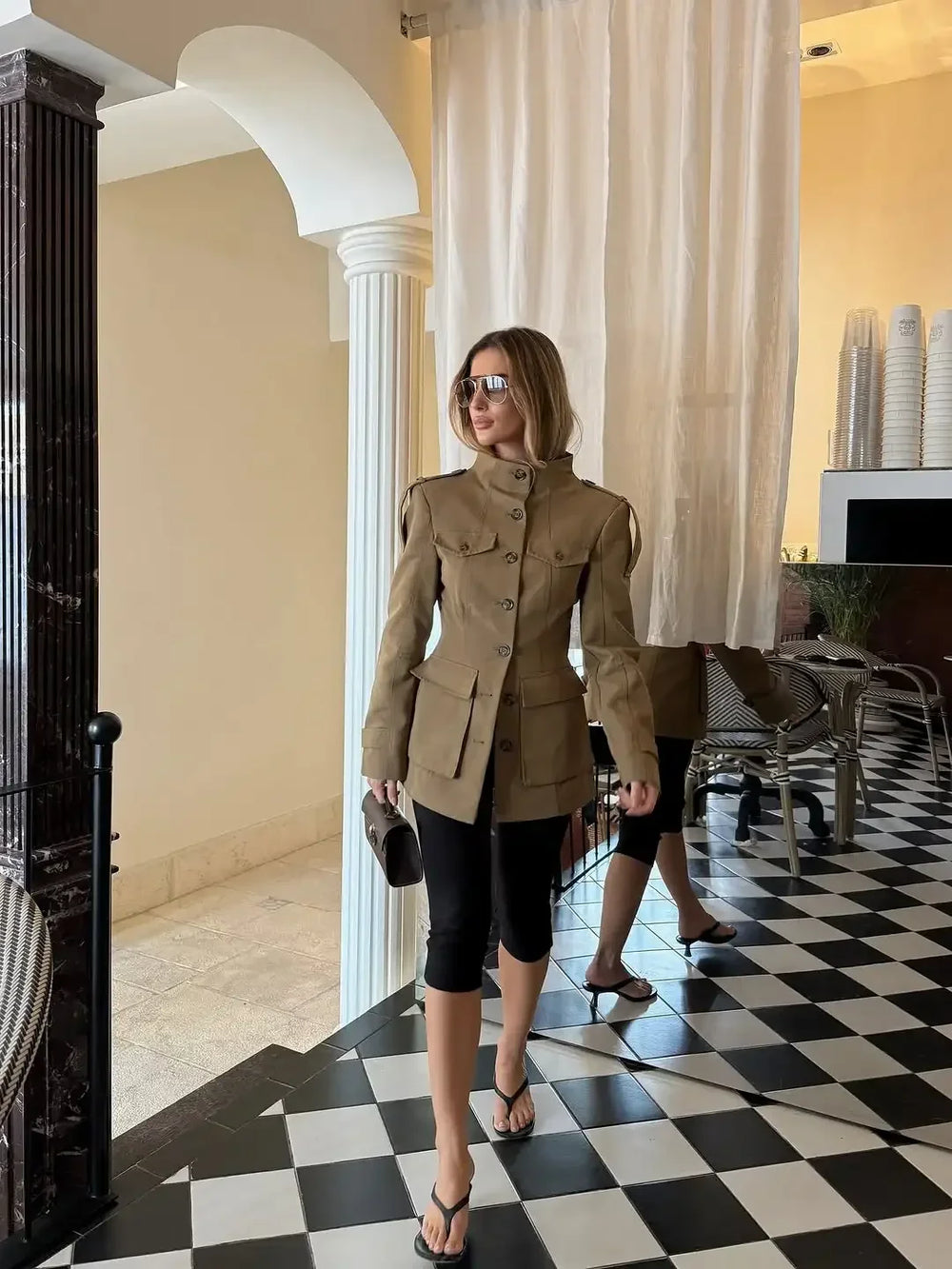 Woman in a beige coat standing in a stylish interior setting with checkered floor and white columns.