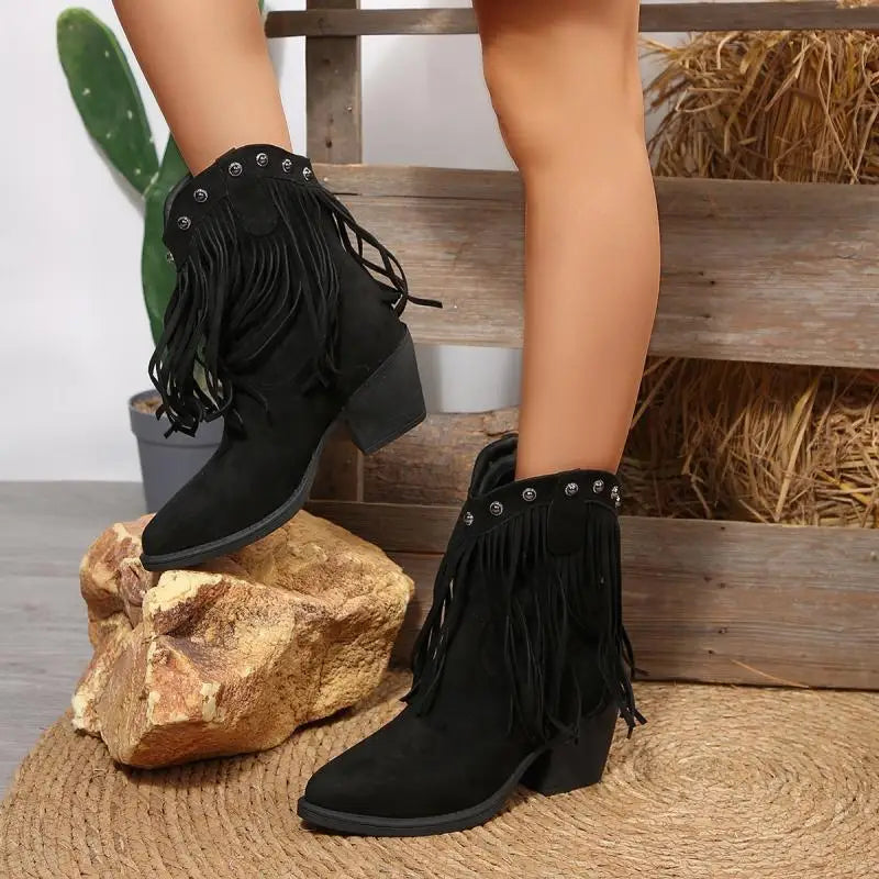 Black fringe boots worn by a person on a rustic background with rocks and hay.