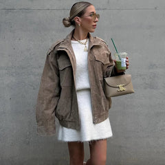 Woman in a brown jacket and white dress holding a drink against a gray wall