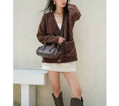 Woman wearing a brown cardigan, white skirt, and black boots holding a black handbag against a light background.