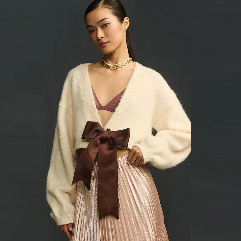 Woman wearing a cream cardigan with a brown bow and pleated skirt against a dark background