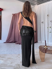 Woman wearing a black sequin dress with a slit in a room with curtains and a table.