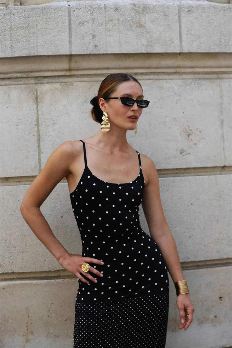 Woman wearing a black polka dot dress with gold accessories against a stone wall.