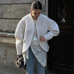 Woman wearing a white coat and blue jeans standing against a textured wall.
