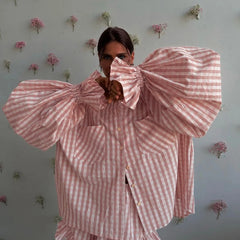 Person wearing a pink and white checkered outfit with large sleeves against a light gray wall with floral decorations.