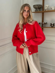Woman wearing a red cardigan over a white shirt and beige pants, standing in a room with decorative shelves.