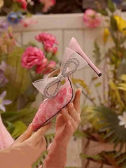 Pink high-heeled shoes with a bow held by a hand against a floral background