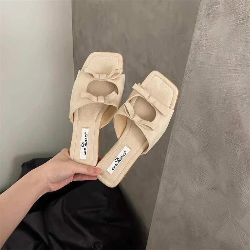 Beige sandals with bow detail held by a hand against a neutral background