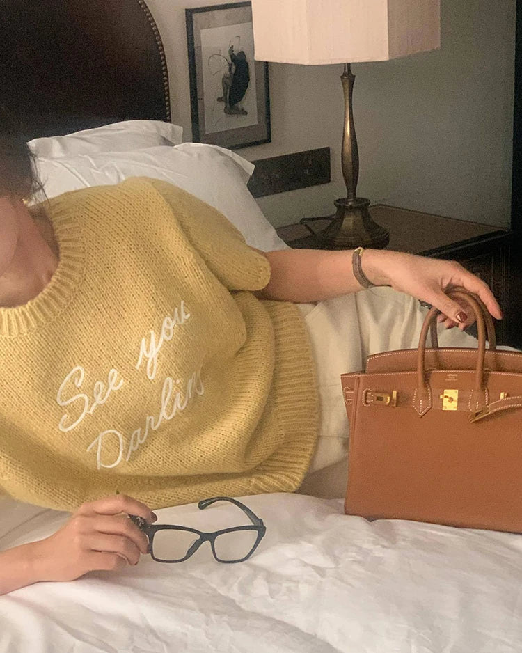 Person wearing a yellow hat with text, holding glasses and a brown handbag on a bed.