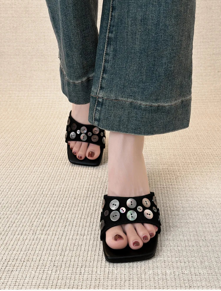 Close-up of feet wearing black square toe wedge heel slide sandals with decorative shell buttons.