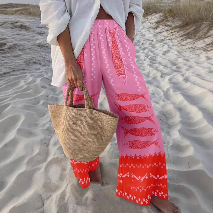 Person wearing pink pants with fish patterns and a white top, holding a straw bag on a sandy beach.