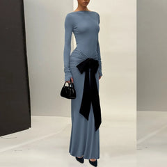 Woman wearing a long blue dress with a black bow and holding a black handbag in a studio setting.