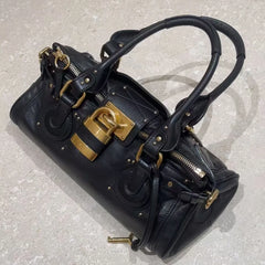 Black leather tote bag with gold lock closure on a textured floor.