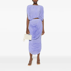 Woman wearing a light purple draped dress holding a white clutch on a white background