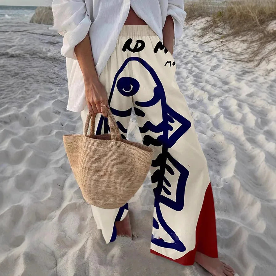 Person on a beach wearing a white outfit with blue and red patterns, holding a woven bag.