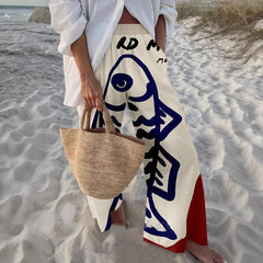 Person on a beach wearing a white outfit with blue and red patterns, holding a woven bag.