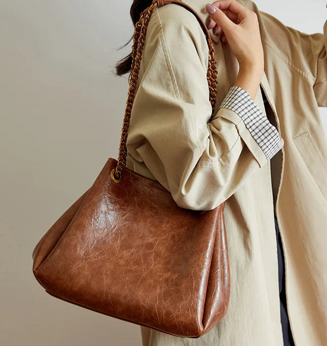 Brown leather handbag held by a person wearing a beige coat against a neutral background