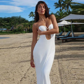 Woman in a white strapless dress standing on a beach with palm trees and lounge chairs in the background.