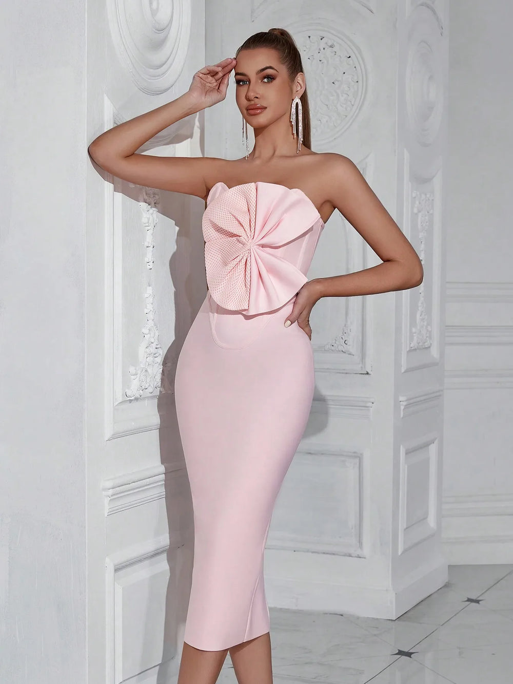 Woman wearing a pink strapless dress with a large bow detail against a white decorative wall.